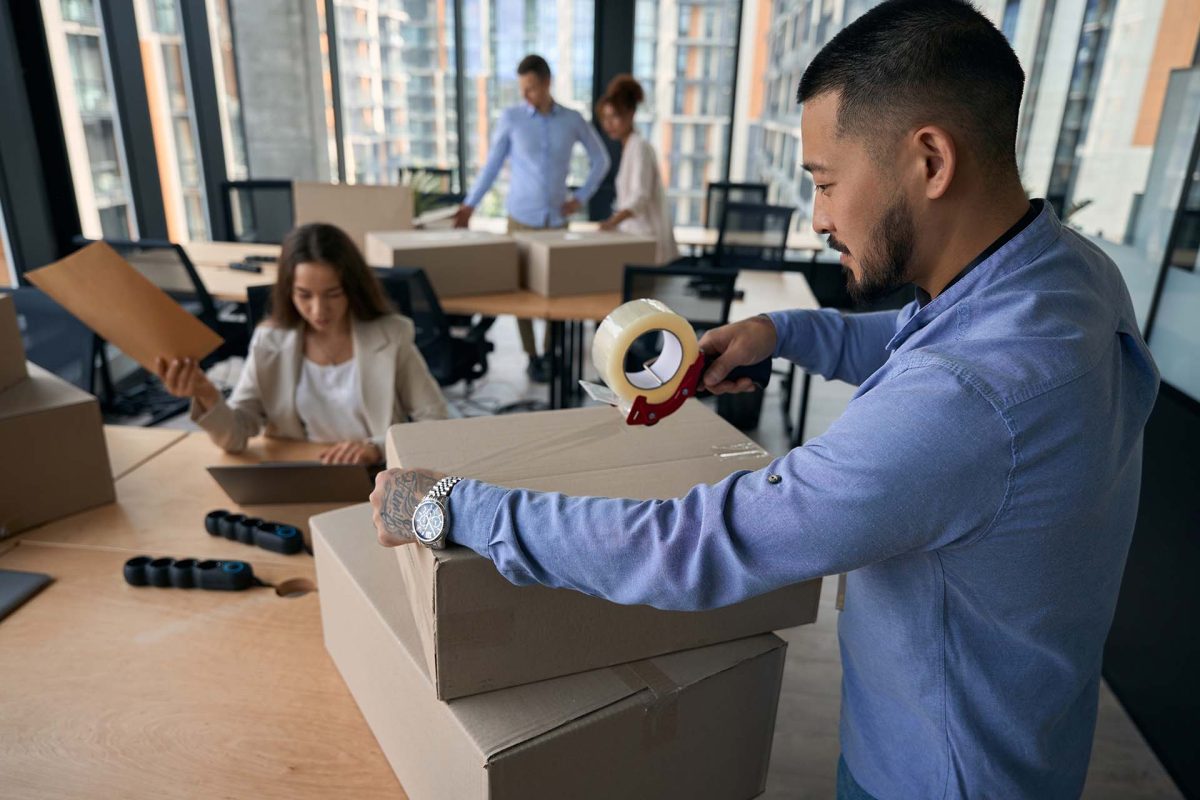 Corporate employee sealing cardboard box with Scotch tape while his female colleague working on laptop with envelope in hand