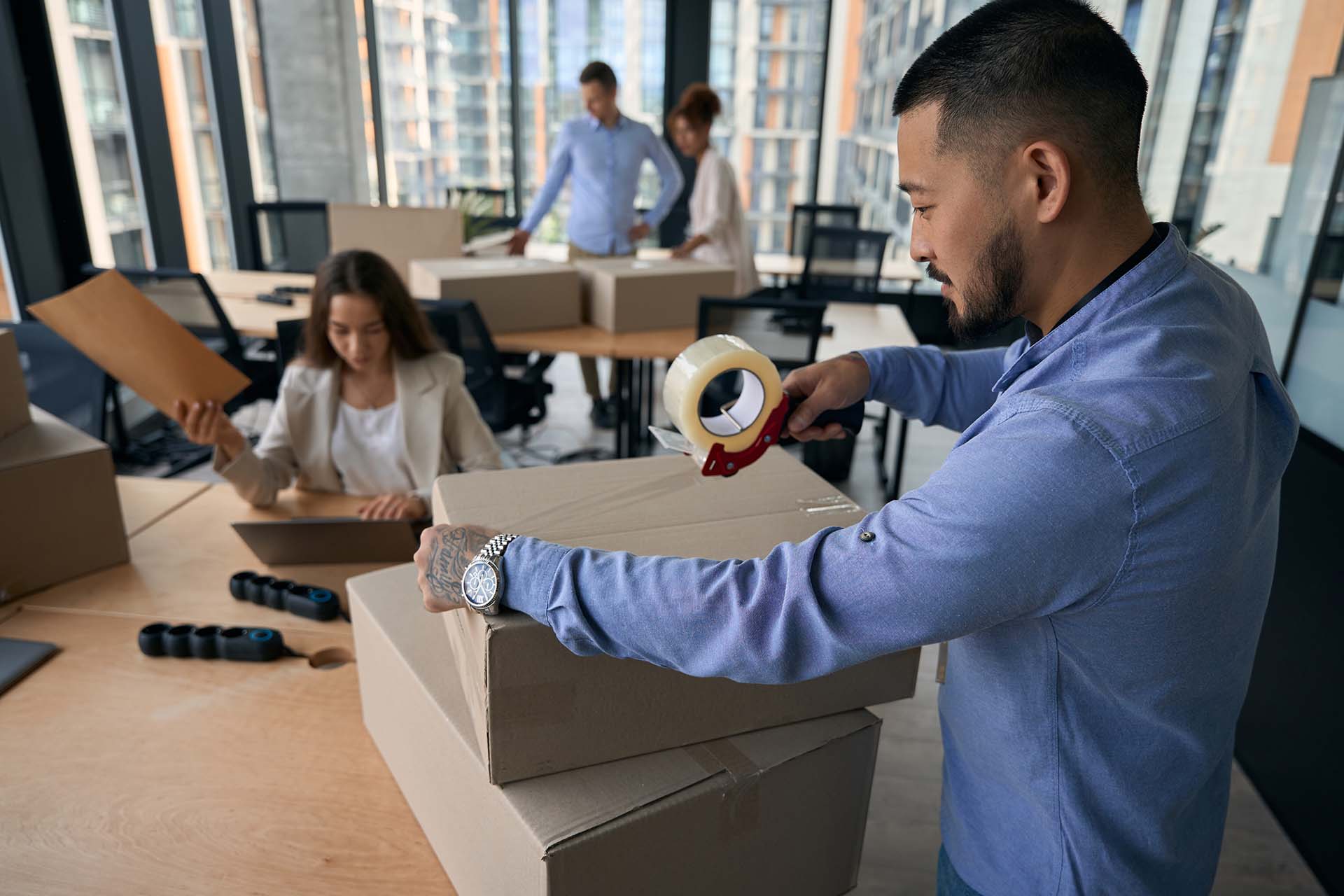 Corporate employee sealing cardboard box with Scotch tape while his female colleague working on laptop with envelope in hand